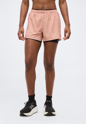 Parte inferiore del corpo di una persona che indossa pantaloncini esterni color pesca sopra pantaloncini aderenti neri, calzini neri e scarpe da ginnastica bianche e nere.