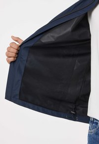 Hand hält offene marineblaue Jacke, zeigt schwarzes Netzfutter im Inneren und verstellbaren Kordelzug am Saum, getragen über weißem Hemd und Jeans.