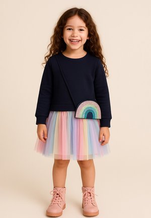 Marineblaues Sweatshirt, pastellfarbener Tüllrock in Regenbogenfarben, pinke Stiefel. Tasche in Form eines Regenbogens mit bunten Pailletten. Lockige Haare, lächelnd.