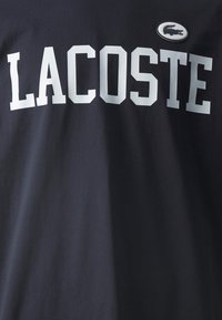 Tummansininen puuvillainen t-paita, jossa on rohkeat valkoiset kirjaimet "LACOSTE" ja pieni brodeerattu krokotiililogo vasemmalla puolella.