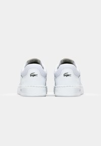Witte leren sneakers met getextureerde achterkant, een zwart krokodillenlogo en details op de hiel, platte rubberen zolen. Minimalistisch ontwerp.