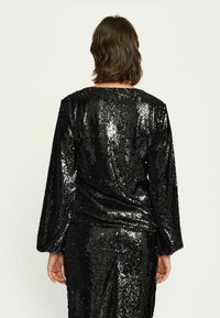 Zwarte pailletten top met lange mouwen, ronde halslijn en een slank, strak ontwerp. Textuur vangt het licht, waardoor een glinsterend effect ontstaat.