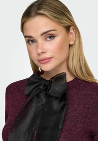 Maglione bordeaux lavorato a maglia con un grande fiocco in raso nero al collo. Texture liscia con un aspetto morbido e peloso.