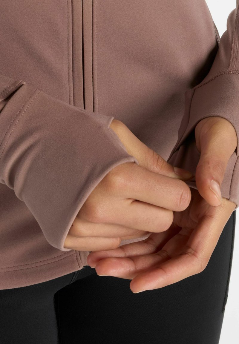 Top de manga larga de corte relajado en suave material malva con agujeros para los pulgares y detalles de costura, combinado con leggins negros. Primer plano de manos ajustando la tela.