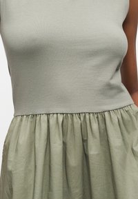 Κοντινό πλάνο αμάνικου φορέματος με εφαρμοστό πλεκτό μπούστο και φουσκωτή, υφασματινή φούστα σε απαλούς αποχρωματισμούς πράσινου φασκόμηλου.