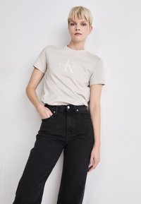 Svetlo béžové bavlnené tričko s okrúhlym výstrihom a krátkymi rukávmi, ktoré má jednoduché biele logo "Calvin Klein Jeans". Skombinované s čiernymi džínsami s vysokým pásom.