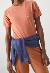 Camiseta de manga corta naranja con un logo de Patagonia. Un suéter azul est á atado alrededor de la cintura. Textura suave, corte relajado, diseño casual.