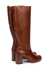 Une paire de bottes en cuir marron hautes avec des talons blocs, une fermeture éclair sur le côté et des détails de broderie décorative près de la cheville, sur fond blanc.