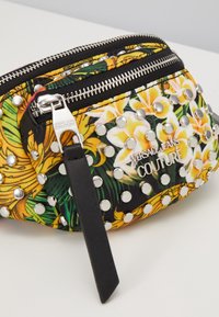 Petit sac Versace Jeans Couture avec imprimé floral jaune, clous argentés, double fermeture éclair et tirette en cuir noire, sur une surface blanche.