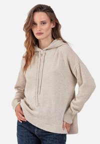 Beige Kapuzenpullover aus weichem Strickstoff mit Kordelzugkapuze, gerippten Bündchen und einem Saum mit vorne kürzerem Verlauf. Kombiniert mit dunkelblauen Jeans.