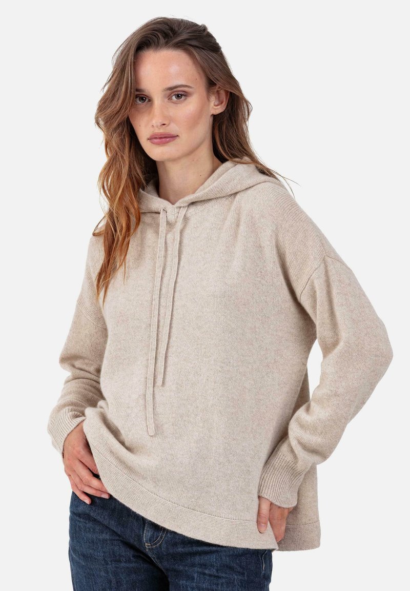 Beige Kapuzenpullover aus weichem Strickstoff mit Kordelzugkapuze, gerippten Bündchen und einem Saum mit vorne kürzerem Verlauf. Kombiniert mit dunkelblauen Jeans.