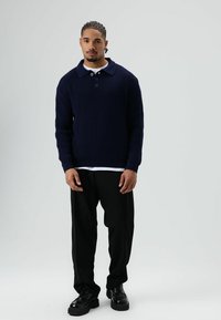 Jonge man met gevlochten haar, gekleed in een donkerblauwe gebreide trui, zwarte broek en zwarte schoenen, staand tegen een effen lichte achtergrond.