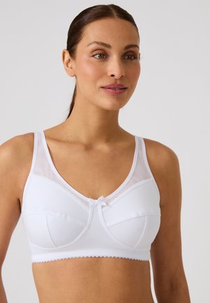 Donna con capelli castani raccolti indossa un reggiseno bianco con coppe morbide, spalline larghe e un piccolo dettaglio a fiocco davanti su uno sfondo semplice.