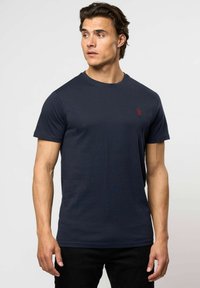 Egy férfi sötétkék, rövid ujjú pólót visel, amelyen egy kis piros hímzett logó található a mellkasán, és balra néz.