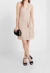 Robe sans manches rose pâle avec un col en V, tissu texturé et taille nouée. Associée à une pochette noire duveteuse et des chaussures à talons hauts à motifs.