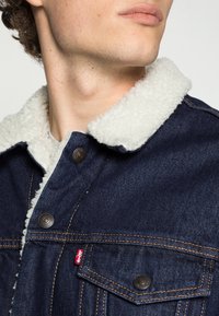 Jeansjacka med en vit, fleece-fodrad krage, kopparfärgade knappar och en liten röd varumärkesetikett på vänster bröstficka.