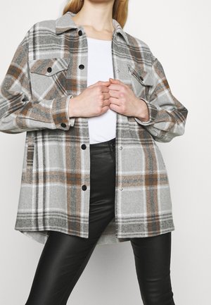 Femme portant une chemise à carreaux grise, marron et blanche sur un haut blanc et un pantalon en cuir noir, les mains jointes sur la poitrine.