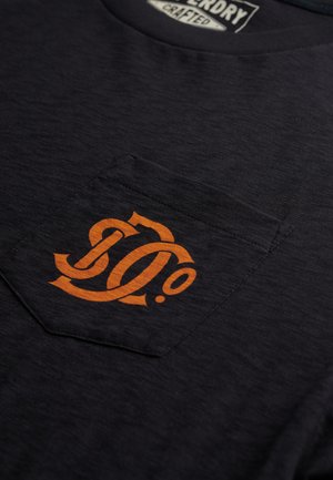 Mørkeblå T-shirt lavet af tekstileret stof med et orange logo på venstre lomme. Udhalsingen er rund, og pasformen er afslappet.