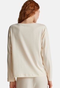 Frau mit roten Haaren, die ein locker sitzendes, langärmliges cremefarbenes Oberteil und passende cremefarbene Ripp-Hosen trägt, von hinten gezeigt.