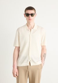 Chemise courte à manches courtes de couleur beige clair avec des rayures verticales, devant boutonné et col. La personne porte également des lunettes de soleil noires et un pantalon beige.