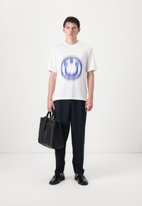 T-shirt en coton blanc avec un graphique bleu, coupe loose. Associé à un pantalon bleu marine, des chaussures en cuir noires et un sac fourre-tout noir structuré.