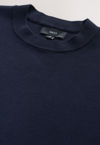 Tumši zils džemperis ar apaļu kaklu, izgatavots no mīkstas teksturētas auduma, ar zīmola "NEXT" nosaukuma etiķeti, kas šūta pie apkakles.