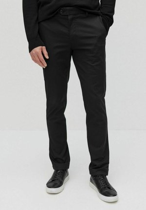 Black trousers in a slim fit, designed with a flat front, side pockets, and soft fabric. Paired with black sneakers featuring white soles.