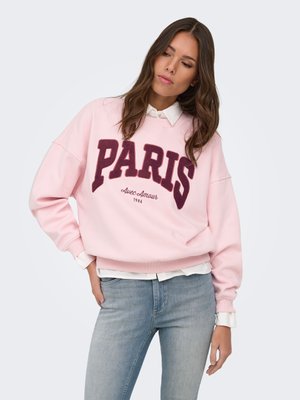 Femme portant un sweat-shirt rose avec « PARIS Avec Amour 1986 » en bordeaux, associé à un jean bleu clair, debout devant un fond uni.