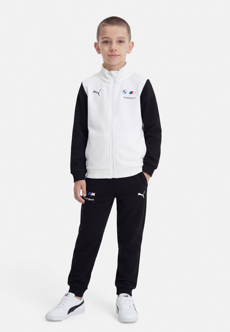 Αγόρι στέκεται φορώντας λευκή και μαύρη φόρμα BMW Motorsport Puma με λογότυπα και λευκά αθλητικά Puma σε λευκό φόντο.