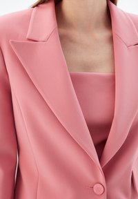 Kobieta nosząca dopasowaną różową marynarkę z klapami w kształcie litery notch, na której ma dopasowany różowy top z kwadratowym dekoltem, odsłaniający dekolt i obojczyk.