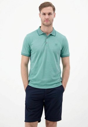 Mann mit kurzen Haaren, der ein türkisfarbenes Poloshirt und marineblaue Shorts trägt, steht mit den Händen in den Taschen vor einem schlichten weißen Hintergrund.