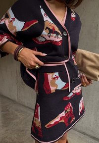 Σετ από μαύρο πλεκτό ζακέτα και φούστα με ροζ λεπτομέρειες, διακοσμημένα με κόκκινα και καφέ σχέδια εμπνευσμένα από ροντεό, χρυσά κοσμήματα και μπεζ τσάντα χειρός.