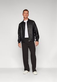 Svart lærjakke med glidelås foran, kombinert med en hvit t-skjorte, svarte bukser og hvite joggesko; modellen står mot en ensfarget bakgrunn.
