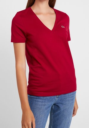 Donna che indossa una maglietta rossa a maniche corte con scollo a V e un piccolo logo bianco sul petto e jeans blu, in piedi davanti a uno sfondo semplice.