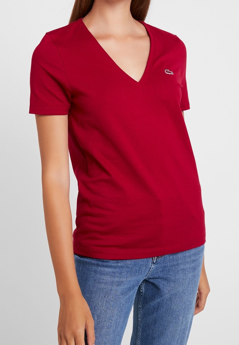Femme portant un t-shirt rouge à manches courtes et col en V avec un petit logo blanc sur la poitrine et un jean bleu, debout devant un fond uni.