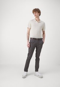 Hellbeige strukturierte Kurzarm-Polohemd, kombiniert mit schmal geschnittenen, dunkelgrauen Hosen und weißen Sneakers. Ein brauner Gürtel sorgt für den Kontrast.