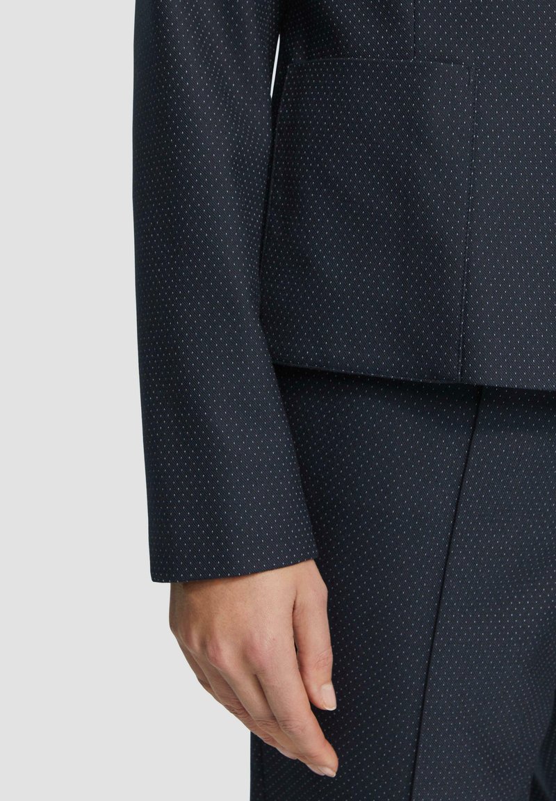 Marineblazer met witte stippen, gestructureerde pasvorm, reverskraag en een enkele borstzak, gemaakt van een zachte stof. Langmouw detail zichtbaar.