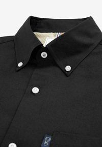 Czarna koszula na guziki z białymi guzikami i zapinanym kołnierzykiem, z małą metką z logo na dolnej, przedniej stronie w pobliżu kieszeni.