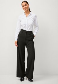 Frau lächelt, trägt ein weißes Hemd mit Knöpfen, das in hoch taillierte schwarze, weit geschnittene Hosen gesteckt ist, dazu schwarze spitze Absätze.