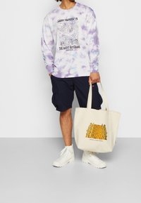 T-shirt à manches longues tie-dye en violet et blanc avec un motif graphique, short cargo bleu marine et sac fourre-tout beige avec un motif d'échiquier.