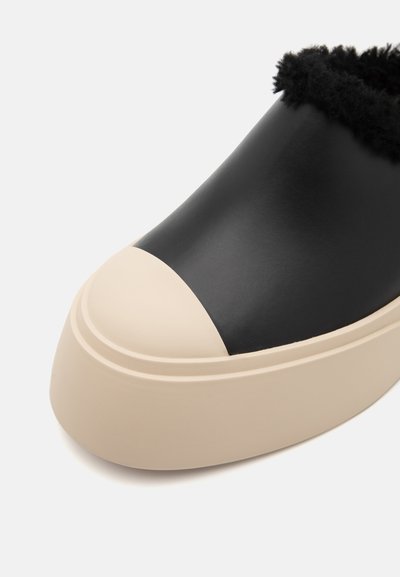 Chaussure noire en cuir à enfiler avec un bout rond en caoutchouc beige et une doublure en fourrure noire texturée, présentant un design épuré et une surface lisse.