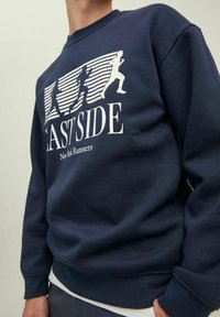 Felpa blu marina con figure in corsa bianche e testo "EAST SIDE New York Runners" indossata da una persona dal collo ai fianchi.