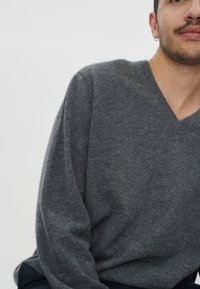 Grauer, leichter V-Ausschnitt-Pullover mit langen Ärmeln, mit glatter Textur und lockerem Schnitt, teilweise von einer Person getragen.