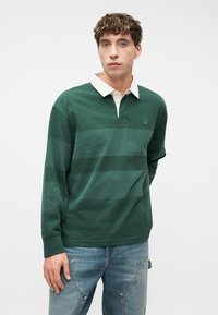 Νεαρός άνδρας με σγουρά μαλλιά, φορώντας σκούρο πράσινο ριγωτό ράγκμπι πουκάμισο με λευκό γιακά και μπλε τζιν, στέκεται μπροστά σε απλό λευκό φόντο.