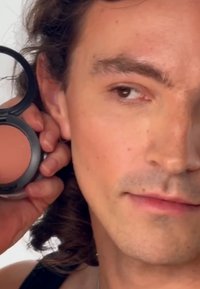 Blush compact dans un container rond noir tenu près du visage. Le produit a une couleur pêche douce. Le modèle a des cheveux ondulés de longueur moyenne.