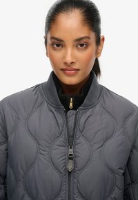 Giacca bomber grigia trapuntata con chiusura a zip, colletto alto e rivestimento interno nero a contrasto. Presenta cuciture curve e un design elegante.
