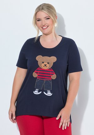 Smiling woman with blonde hair wears a navy shirt with a teddy bear in jeans and a red striped shirt, paired with red pants.