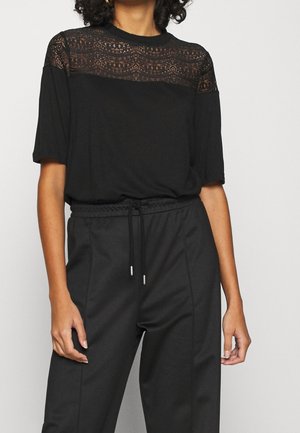 Femme portant un haut noir à manches courtes avec un détail en dentelle sur le haut de la poitrine et un pantalon noir taille haute avec cordon à la taille.