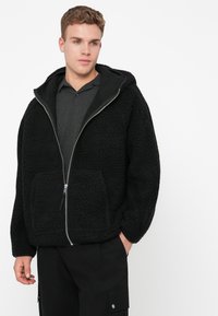 Casaco de fleece preto com uma superfície espessa e texturada, fecho zip na frente e capuz. Apresenta dois bolsos frontais e um corte descontraído.