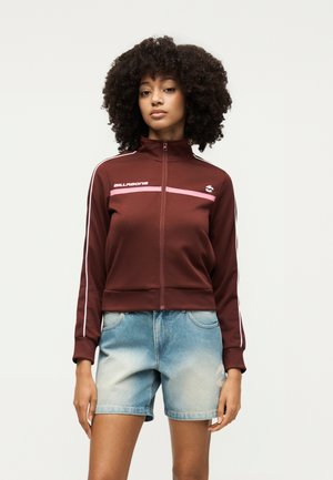 Donna con capelli ricci che indossa una giacca bordeaux con zip e strisce rosa e pantaloncini di jeans azzurri, in piedi davanti a uno sfondo semplice.